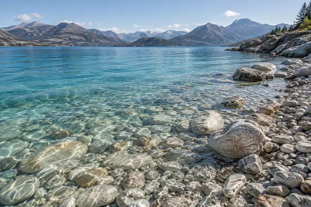 AI illustration: Clear mountain-lake water over rounded pebbles, with shore grasses and dry slopes in the distance.