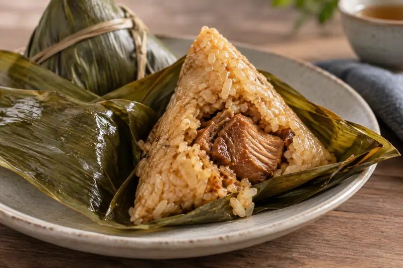 AI illustration: A natural close-up food photograph of a pork zongzi partly unwrapped from glossy green bamboo leaves.