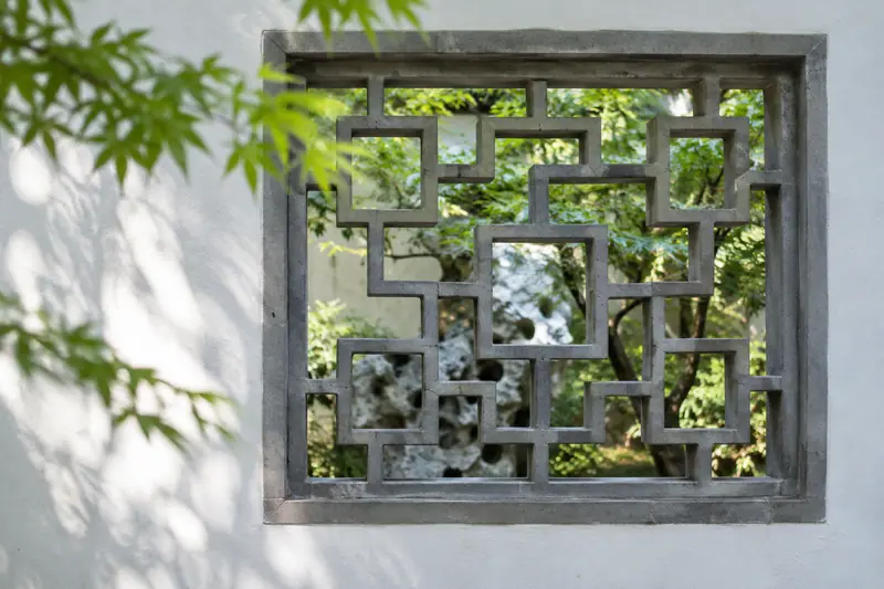 AI illustration: A grey geometric lattice window in a white garden wall, with leaves and a natural stone visible beyond.