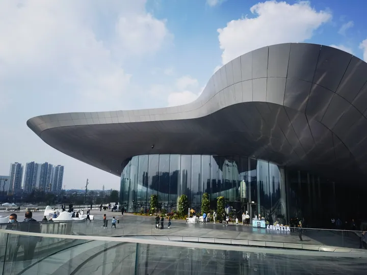 The Chengdu Science Fiction Museum's exterior: a broad silver-grey building with a sweeping curved cantilevered roof over a tall glass curtain wall, with people and model figures on the paved plaza in front, high-rise towers at the left and a blue sky with white clouds.