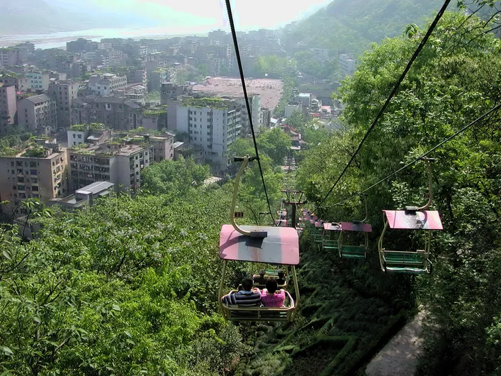 A view from a chairlift on a wooded hillside: a dense green forested slope with a footpath below, pink-roofed and green chairlift cars carrying passengers on cables crossing the frame, and a city of high-rise blocks along the Yangtze in the upper left.