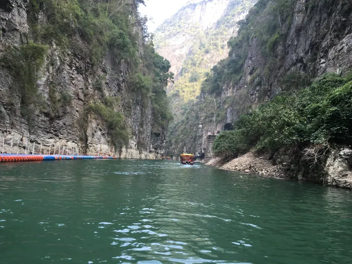 A narrow gorge seen from water level: sheer grey rock cliffs with clinging vegetation on both sides, deep green water across the foreground, a small covered tour boat ahead with passengers, and a line of blue and red floating pontoons along the left bank.