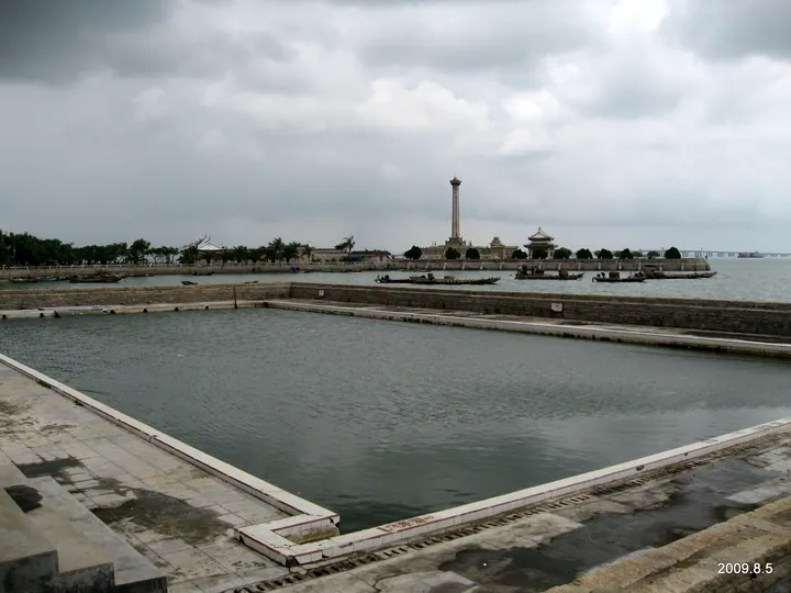 A view across water toward 鳌园: a large rectangular stone-edged water basin with steps in the foreground, a stone causeway with moored boats beyond it, the tall column of the 集美解放纪念碑 with a pavilion beside it on the far shore, trees and a distant bridge under heavy grey cloud.