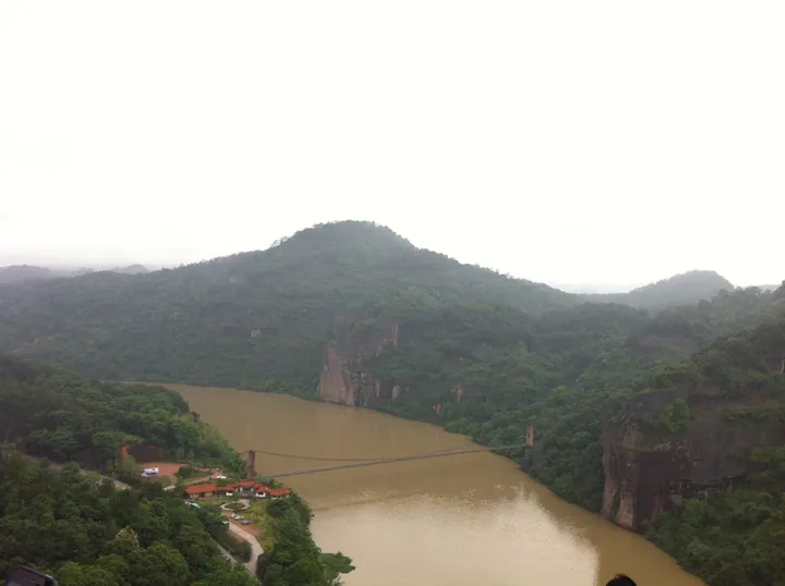 An elevated view of a river gorge: a broad muddy-brown river curving between forested hills and reddish sandstone cliffs, a slender suspension footbridge crossing the water, and a visitor area with red-roofed buildings, a road and parked cars on the near bank under a pale overcast sky.