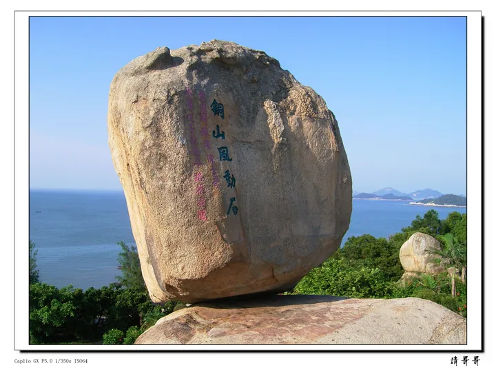 A huge rounded granite boulder balanced on a flat rock ledge, carrying carved characters 銅山風動石 and smaller red inscriptions, with green trees and shrubs around it and blue sea, headlands and distant islands behind; the file has a thin white border with a Caplio GX exposure caption at the lower left and a 請哥哥 watermark at the lower right.