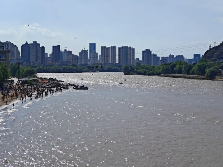A wide view of the Yellow River at Lanzhou with sunlit water across the frame, crowds of bathers and small boats along the near bank at the left, tree-lined banks, and a skyline of high-rise blocks on the far side under a hazy sky with birds overhead.