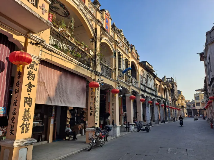 A street of two-storey arcaded shophouses with cream walls, arched verandas and ironwork balconies, hung with red lanterns; vertical signs read 風物舍·陶藝館, 明商旅宿 and 新會土地..., a motorcycle is parked at the kerb, a scooter rider and a seated shopkeeper are the only people, and the street runs away to more arcaded buildings under a clear blue sky.