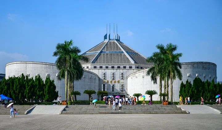 A wide frontal view of a large museum: a low white-tiled building with curved wings and a central pyramid of glass and steel panels topped by three fins, its entrance inscribed 海战博物馆, with a broad flight of steps, palms and a crowd of visitors in the foreground.