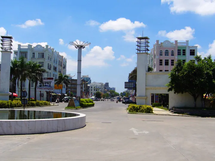 A wide street junction in bright sun: white and pastel arcaded buildings with arched windows and balconies on both sides, palms and clipped shrubs, a marble-rimmed pool in the left foreground, an ornamental lamp column and a traffic-filled street beyond; deep blue sky with cumulus.