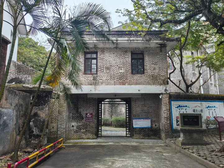A two-storey grey brick house seen frontally: an arched carriage entrance under a flat lintel, two shuttered windows above, a stone plaque reading 陈公馆 on the right, a blue information board and a banner on the boundary wall; palms and a dark tree overhead.