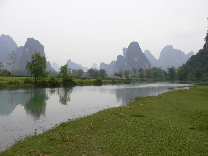 A calm river curving through flat green fields, a small thatched pavilion on the far bank, and a long rank of steep karst peaks receding into haze behind; pale overcast sky and trees along the banks.