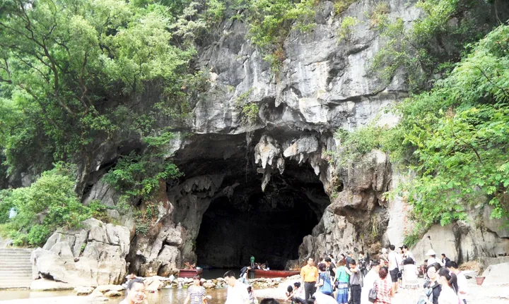 A huge cave mouth opening in a grey limestone cliff, stalactites hanging from its roof and a boat and figures at the dark entrance; dense green trees and shrubs clothe the rock above and beside it, with visitors on a stone stairway and beach below.