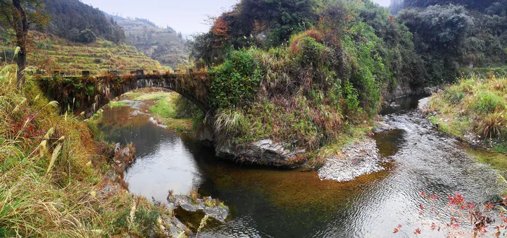 A stone arch bridge with a grassy parapet crossing a shallow rocky stream, terraced fields rising on the hillside behind it, autumn grasses and shrubs along the banks and scattered village buildings on the upper slope under a pale sky.