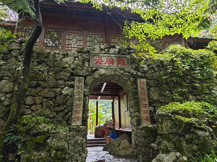 A rough stone archway set into a boulder wall, its keystone panel inscribed 龍騰堡 in red with carved couplets on either side, opening onto a timber gallery with lattice windows; moss, ferns and a large tree frame it, and a child sits at the threshold.