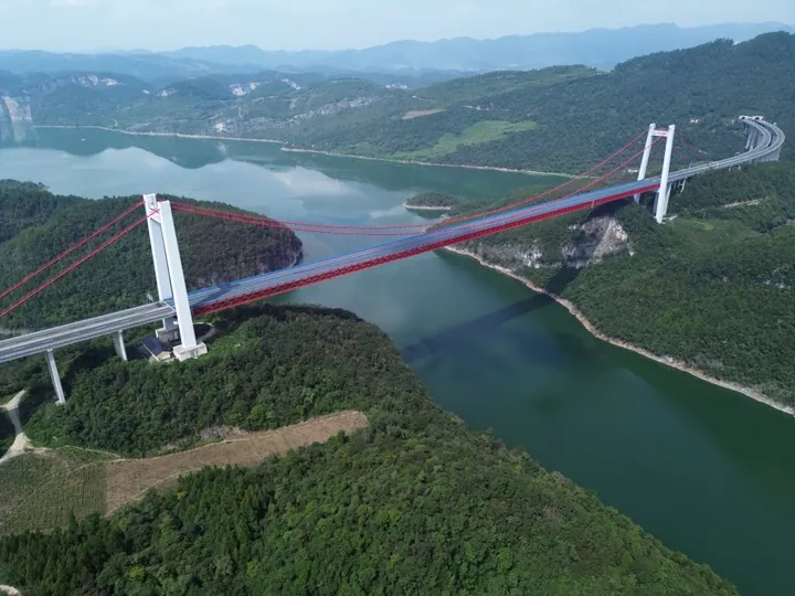 An aerial view of a broad green reservoir among forested karst hills, spanned by a suspension bridge with white pylons and red cables carrying a viaduct; terraced fields and a cliff face at the left and hazy hills beyond.