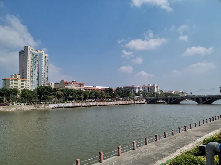 A riverside view of a small town: a broad green river behind a stone-edged promenade with a chained railing, a white and blue multi-storey hotel tower and lower blocks among trees on the far bank, a concrete road bridge crossing at the right, under a blue sky with cumulus.