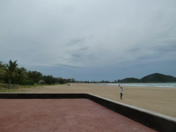 A broad sweep of pale sand beach with a few distant bathers, a headland covered in dark green scrub at the right and a line of coconut palms and low resort buildings at the left, seen past a large dark red paved terrace with black coping in the foreground under an overcast grey sky.