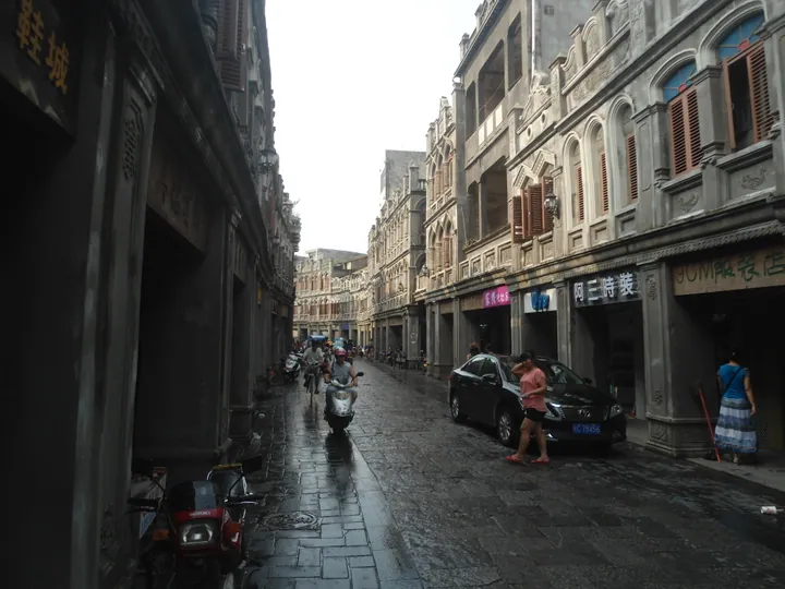 A narrow paved street of two- and three-storey arcaded shophouses in grey weathered stucco with arched windows and louvred shutters and shopfronts (阿三時裝, 90M服裝店); scooters and a black saloon stand in the street, a woman in a pink top walks past, and a deeply shadowed colonnade runs down the left side.