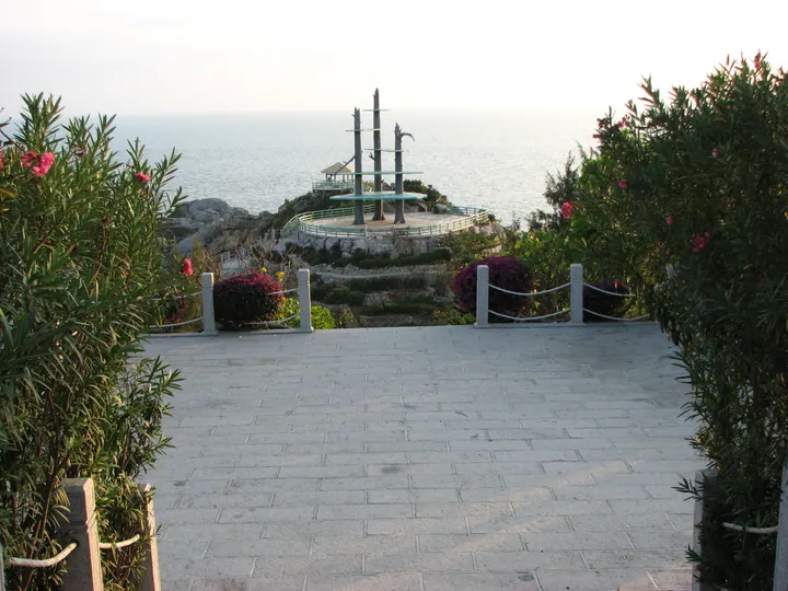 A paved clifftop overlook edged with oleander bushes, looking past a rope railing to a rocky headland carrying a small pavilion and three tall carved timber masts, with open sea and a pale sky behind.