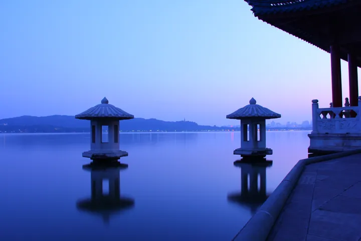 A blue-hour view over glassy West Lake: two stone lantern pagodas standing in the water with mirrored reflections, a moon-viewing terrace and pavilion eave at the right, and the city skyline and hills faint on the far shore.