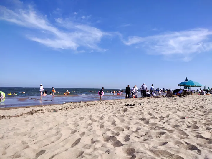 A broad sandy beach with bathers and a blue parasol along the waterline, calm sea behind and a blue sky streaked with cirrus.