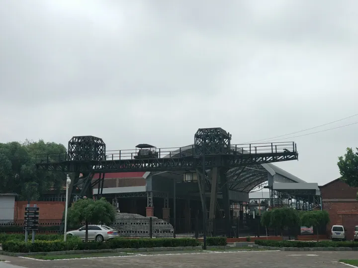 A large brown steel gantry with two lattice towers spanning a trackbed in front of an industrial hall with an arched roof and brick outbuildings, with a hedge, fence, parked car and minibus in the foreground under a blank grey sky.