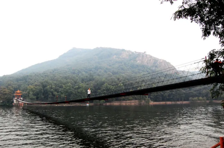 A long suspension footbridge crossing grey water toward a forested hill, with a small pavilion at its far end, a single walker mid-span, willow foliage at the right edge and a hazy white sky.