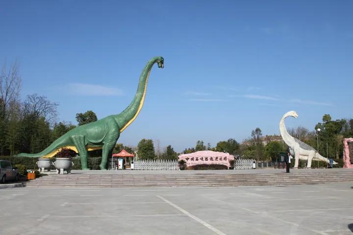 A sunlit paved plaza at the entrance of the Xixia Dinosaur Relics Park: a huge green sauropod statue with a raised neck on the left and a white one on the right, a pink stone wall lettered 中国西峡恐龙遗迹园 between them, a white picket fence, trees and a blue sky; a parked car and a kiosk sit at the left edge.