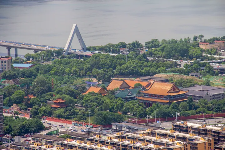 An elevated view over Changsha: the walled Kaifu Temple compound at the centre with several large halls under orange-yellow tiled roofs, a red perimeter wall and dense trees, the broad grey Xiang River behind with a cable-stayed bridge crossing at the upper left, and blocks of flats in the foreground.
