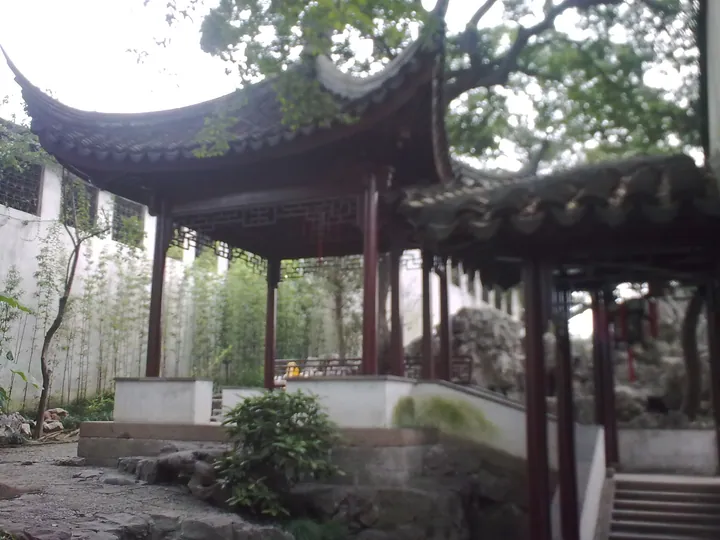 A Suzhou garden courtyard: a square pavilion with upturned grey-tiled eaves and dark timber columns raised on a white-walled stone terrace, a covered walkway with a further eave at the right, bamboo and whitewashed walls behind, and rough rockery boulders and low shrubs along the gravel below.