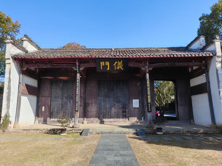 The 仪门 of a Chinese county yamen: a timber gate hall with a grey tiled roof and a black plaque lettered 儀門 in gold above three bays of dark wooden doors, painted couplets on the posts, white side walls, potted plants on the stone step and a slab path crossing a dry grass court to the gate.