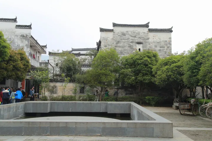 A paved village square in Wuyuan: white-walled Hui-style houses with grey tiled roofs and stepped horse-head gables behind a sunken rectangular stone-walled pool, a large tree in front, a group of visitors at the left, a tricycle and parked bicycles at the right, under a blank pale sky.