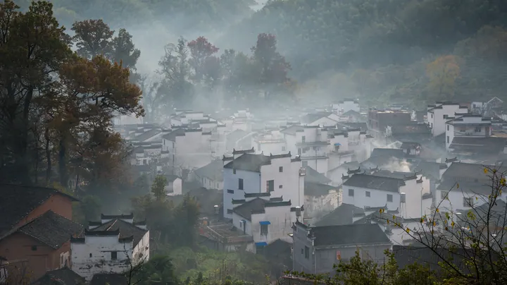 A hillside village of white-walled, grey-roofed houses packed into a wooded valley, autumn foliage in red and orange among the trees, mist or wood smoke drifting between the roofs, a few small modern buildings at the left and a wooded ridge behind.