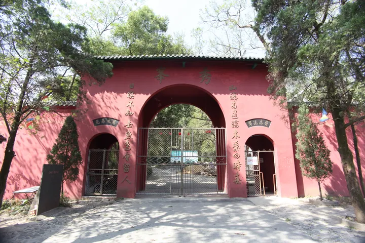 A red-painted entrance gate: a three-arched wall under a grey tiled roof, the central arch lettered 秀峰 in gold with a couplet down each pier, side arches labelled 门票处, iron grilles closing the openings, a dark stone stele on the ground at the left and mature trees at both sides.