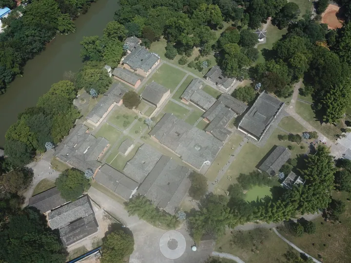 An aerial view of the Yeping revolutionary site: a large complex of grey-tiled single-storey halls and courtyards laid out among lawns and mature trees, with paths, a circular paved plaza with a monument at the bottom, a river and further houses along the upper left and a dirt bank at the upper right.