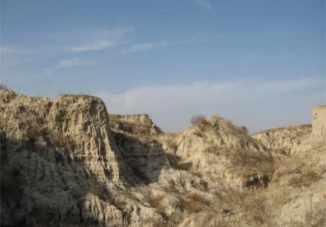 Eroded badland pillars of pale loess rising as a ridge across the frame, their flanks cut into gullies and fluted ribs, with dry golden grass and scrub on the slopes, more eroded ground behind and a clear blue sky with a thin cirrus wisp.