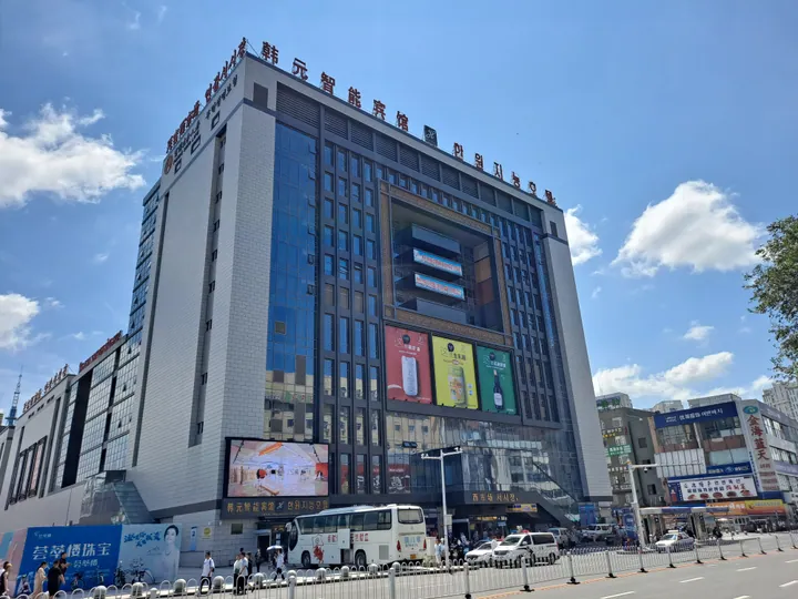 A tall multi-storey market building of white stone and dark glazing seen across its forecourt, carrying the roof sign 韩元智能宾馆 and the vertical signs 서시장 / 西市场 with a large LED screen and advertising panels on its flank; buses, cars and pedestrians along the road in front, other commercial blocks at the right, under a blue sky with cumulus.