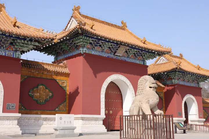 The front gate of the Zhao Mausoleum: a red wall with three arched openings under golden glazed roofs with painted bracket sets, red studded doors, a white marble plaque lettered 清昭陵 on a pedestal at the left, a carved stone lion behind a low railing at the right and an 出口 EXIT sign at the far left, against a clear blue sky.