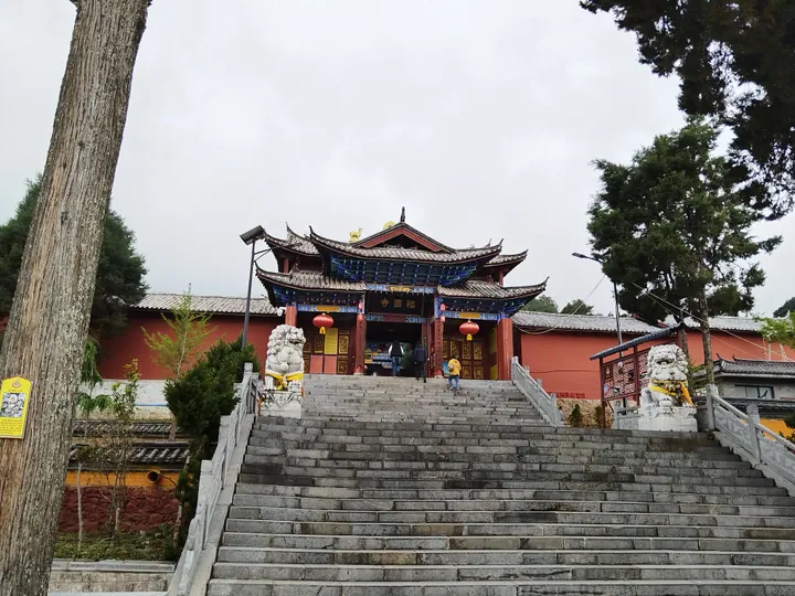 A temple front seen up a broad flight of grey stone steps with carved balustrades: a single-storey hall on a raised platform with a grey tiled roof, dark blue painted eaves and bracket sets, a central plaque lettered 福国寺 above red doors flanked by yellow panels, two white stone lions with yellow ribbons on the steps, two red lanterns under the eaves, red walls with grey capping to either side, a large conifer at the left and a display board on a pole at the far left.