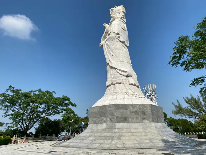 A tall white stone statue of a robed, crowned A-Ma seen from a low angle against a deep blue sky, standing on a grey stone plinth and a stepped octagonal base of pale stone blocks; a paved plaza with a low wall and railings at the bottom left with a seated person and a parked scooter, leafy green trees at both sides and a lattice telecommunications mast behind the statue's shoulder.