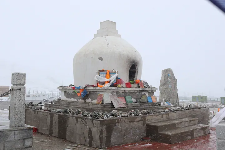 A whitewashed stupa with a stepped square finial standing on a square stone platform over a stepped base, draped with white scarves and hung with coloured prayer flags, with offerings, small plaques and a dark doorway at its right; a carved stone stele with prayer flags stands beside it, chains and stone posts cross the foreground, snow lies on the ground and road, and cars and a line of prayer flags are just visible in the white haze behind.