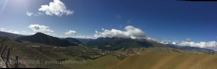 A wide mountain panorama: rounded green and dry-gold slopes with terracing sweeping down in the foreground, a valley floor with a small town, fields and a road in the middle distance, darker forested ridges beyond and a snow-streaked range on the horizon; a deep blue sky with cumulus and cirrus, a dark hill at the left, a fence post at the left edge, a small white structure on the right ridge and a white watermark © 郭泽泽泽泽泽 / guozzzzz.lofter.com at the lower left.