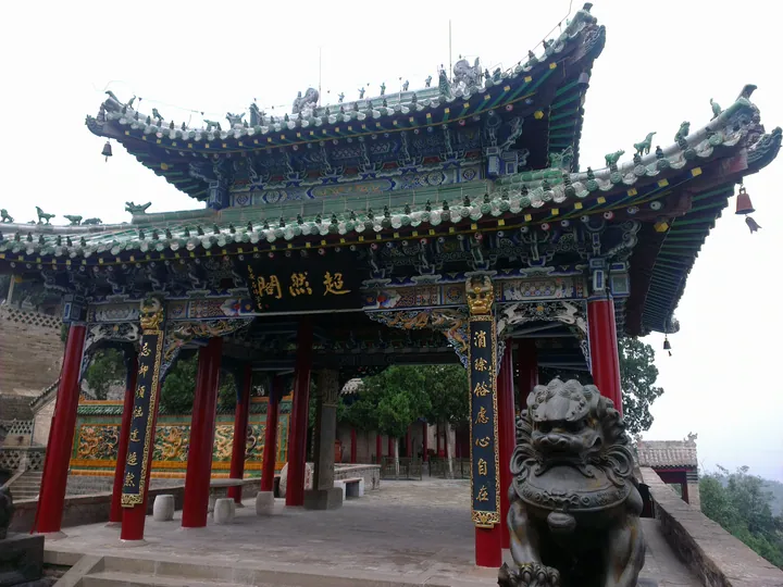 An ornate temple pavilion in the frame centre: a double-eaved roof of dark green glazed tiles with rows of small ridge figures, bells hanging from the corner eaves, painted beams in blue, green and gold, four red columns carrying a pair of gilt-lettered couplets, and a black plaque lettered 超然亭 in gold above the opening; a carved stone lion on a plinth at the lower right, glazed-tile screen walls and a staircase behind at the left, and a hillside with trees beyond under a flat pale sky.