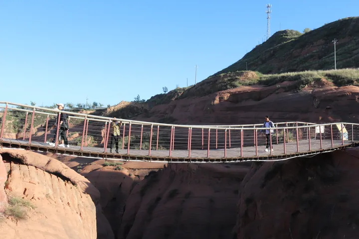 A suspension footbridge of steel posts, timber deck and pale rails curving across the frame from left to right with three visitors walking on it; below and behind it the walls of a narrow canyon of red sandstone cut into horizontal wavy strata, with a dry gully floor, sparse green shrubs on the ledges, a grassy slope rising at the right, a lattice mast and a lamp post on the ridge and a clear blue sky.