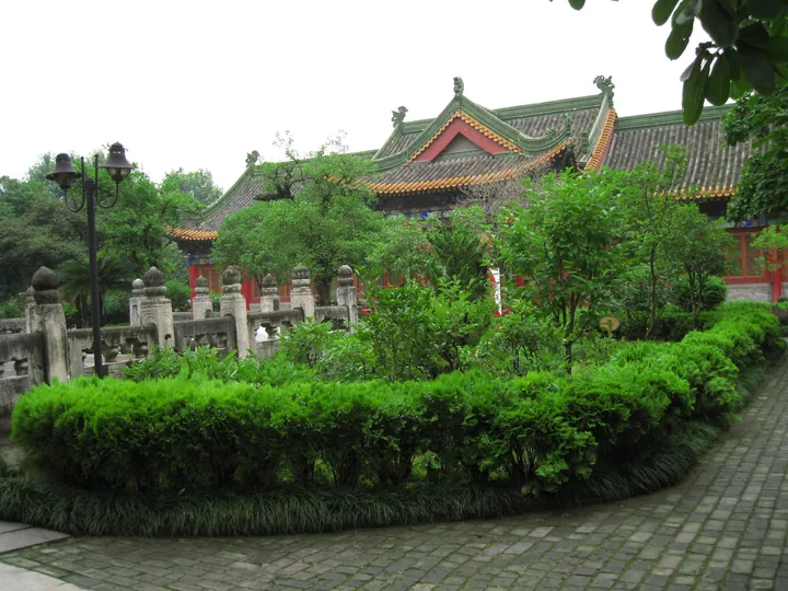 A courtyard of the 古汉台 complex: a traditional hall with a grey tiled roof, green glazed ridge tiles and an ornate finial, red doors and a red banner, seen across a clipped box hedge and a lawn with trees; a carved stone balustrade with lion-topped posts runs along the left, a black double lamp post stands in front of it, and a paved path of grey bricks curves away at the right under a bright overcast sky.
