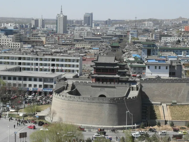 An elevated view over 榆林's old town: a long grey brick city wall with a crenellated parapet and a bastion sweeping across the middle distance, a gate tower with a double-eaved grey roof rising behind it, a street of traditional roofs with further tower-like structures receding into the town, modern apartment blocks and office towers around the edges, a road with cars, pedestrians and blossom trees at the lower left and a wooded riverbank at the right under a clear pale sky.