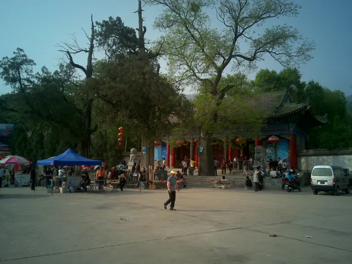 The entrance front of 周公庙: a hall with red columns, blue-and-gold painted brackets and a green tiled roof behind two stone lions, red lanterns strung along it, a paved plaza in front with market tents and visitors, and tall trees rising behind the roofs.