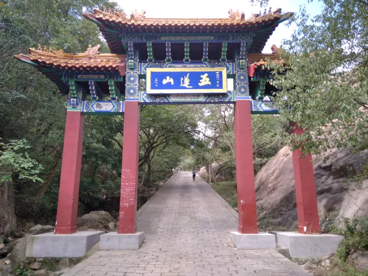 The 五莲山 entrance paifang: four red piers on granite bases carrying a painted blue-and-green beam with a blue plaque lettered 五莲山 in gold, under a tiled roof with ridge figures, straddling a paved stone path that runs into wooded, boulder-strewn hills.