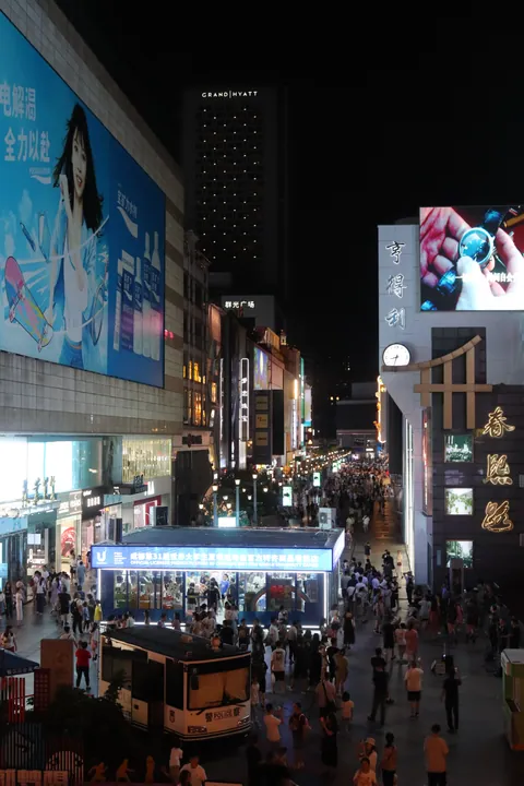 A night view down 春熙路: a packed pedestrian street between tall buildings hung with huge illuminated advertising screens, the 亨得利 clock sign and 春熙路 lettering at the right, the Grand Hyatt tower beyond, and crowds and a police van on the paving.