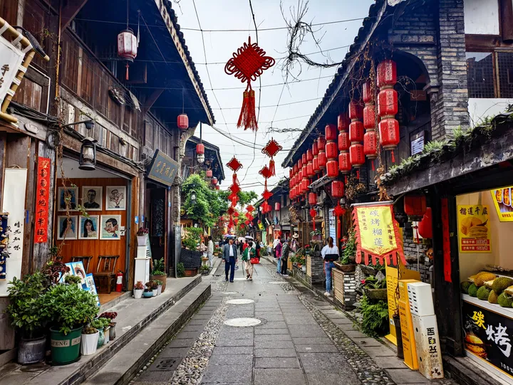 A narrow stone-paved street in the 灌县古城 old town: dark timber shopfronts on both sides, rows of red lanterns strung overhead and along the eaves, framed photographs and plants at the left, and visitors walking away down the lane.